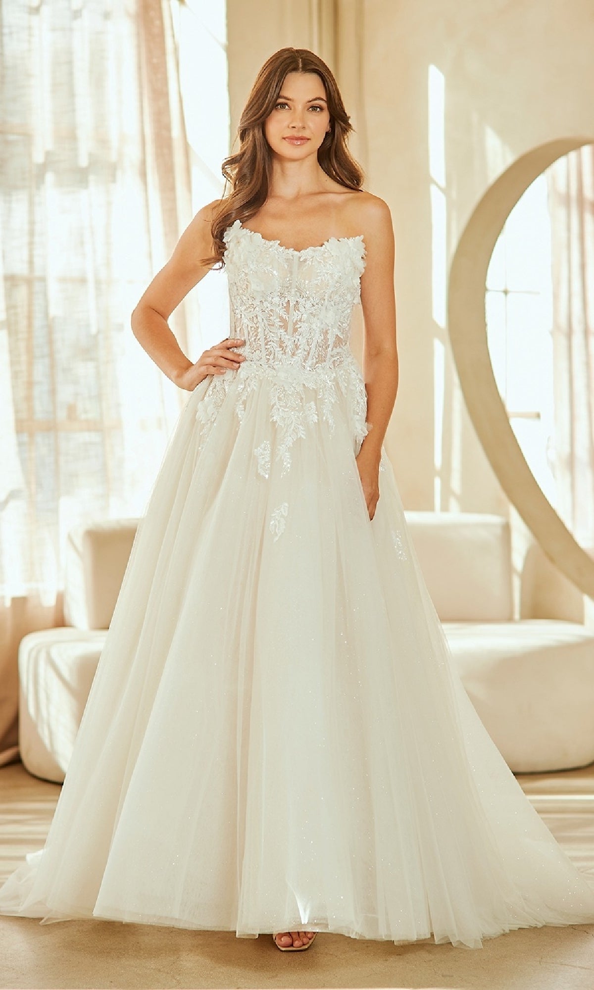 A woman in the Amelia Couture 2141 long wedding dress, a strapless ball gown with floral embellishments, poses indoors with one hand on her hip before a sofa and large decorative mirror.
